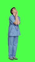 Side view Hospital nurse worshipping God by prayer against greenscreen backdrop, praying to jesus christ and having hope in christianity and spirituality. Medical assistant doing meditation. Camera A. video