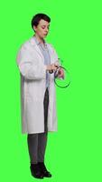 Side view General practitioner with a white coat using stethoscope in studio, preparing to examine patients at checkup appointments. Female doctor in uniform standing against greenscreen backdrop. Camera A. video