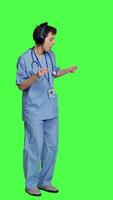 Side view Nurse having fun listening to music on wireless headphones, wearing scrubs and dancing around against greenscreen backdrop. Medical assistant showing groovy funky dance moves. Camera A. video