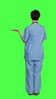 Back view Medical assistant pointing at something aside and doing web commercial, creating advertisement against greenscreen backdrop. Young nurse with scrubs showing a thing to left or right sides. Camera A. video