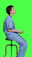 Profile Nurse browsing online websites on smartphone app in studio, sitting on a chair against greenscreen backdrop. Medical specialist in scrubs using mobile phone and social media texting. Camera A. video