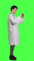 Profile General practitioner playing video games on mobile phone app, having fun with online gaming competition against greenscreen backdrop. Cheerful doctor relaxing with internet game. Camera A.