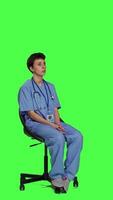 Side view Medical assistant is waiting for patients to attend exam appointments, feeling impatient while she sits in a chair. Nurse with stethoscope and scrubs waits for people against greenscreen. Camera A. video