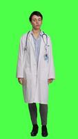 Front view Physician saying no and showing negative reaction symbol in studio, wearing a white coat and standing against greenscreen backdrop. Woman medic feeling displeased, rejection sign. Camera A. video