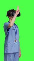 Side view Medical assistant working with virtual reality glasses against greenscreen backdrop, using interactive 3d vision on vr headset to do checkup examination. Young nurse uses modern gadget. Camera B. video