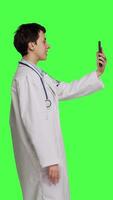 Profile Medic specialist having remote videocall conversation with patients on smartphone internet connection, greenscreen backdrop. Doctor discusses about disease with people on videoconference. Camera B. video