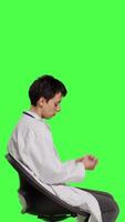 Profile Physician in white coat waiting for patients at consultations, feeling impatient sitting on a chair against greenscreen backdrop. Medic practitioner with stethoscope waits for people. Camera B. video