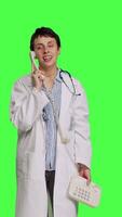 Front view Physician answering landline phone call against greenscreen backdrop, talking to patients about checkup appointments. Medic having remote discussion on retro telephone with cord. Camera B. video