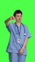 Front view Displeased medical assistant shows thumbs down in studio, presenting her disapproval and disagreement with dislike symbol against greenscreen. Nurse feeling unhappy with rejection sign. Camera B. video