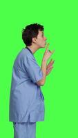 Profile Medical assistant asking to keep quiet and be silent against greenscreen, showing hush mute symbol to keep privacy and silence. Young nurse showing secrecy taboo sign. Camera B. video
