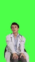 Front view Physician in white coat waiting for patients at consultations, feeling impatient sitting on a chair against greenscreen backdrop. Medic practitioner with stethoscope waits for people. Camera B. video