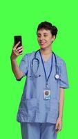 Front view Nurse answering videocall meeting connection on work break, using smartphone to connect with friends on online videoconference chat. Specialist in scrubs stands against greenscreen. Camera B. video