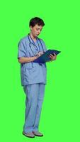 Side view Confident hospital nurse taking notes for the next medic appointment, standing over greenscreen backdrop. Medical assistant with uniform writing patient information on clipboard papers. Camera A. video