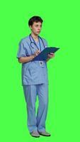 Side view Medical assistant writing checkup information on clipboard files, taking notes and making doctor appointments with checklist. Nurse wearing blue scrubs stands against greenscreen backdrop. Camera A. video