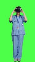 Front view Medical assistant uses interactive vr glasses to check disease treatment, looking at patient examination files with virtual reality headset and lens. Nurse against greenscreen backdrop. Camera A. video