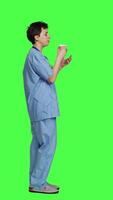 Profile Healthcare specialist enjoying hot coffee cup against greenscreen backdrop, drinking caffeine refreshment in blue scrubs. Medical assistant serving beverage at work, friendly staff. Camera A. video