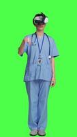 Front view Health specialist using vr headset to examine patient diagnosis, stands against greenscreen backdrop. Nurse works with artificial intelligence virtual reality glasses, three dimensional. Camera A. video