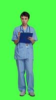 Front view Medical assistant writing checkup information on clipboard files, taking notes and making doctor appointments with checklist. Nurse wearing blue scrubs stands against greenscreen backdrop. Camera A. video