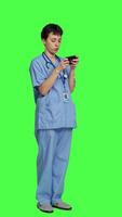 Side view Medical assistant playing mobile video games on smartphone app, taking a break from healthcare work. Nurse enjoys gaming tournament on phone, standing against greenscreen backdrop. Camera A.