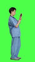 Profile Medical assistant playing mobile video games on smartphone app, taking a break from healthcare work. Nurse enjoys gaming tournament on phone, standing against greenscreen backdrop. Camera A.