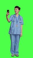 Front view Nurse answering videocall meeting connection on work break, using smartphone to connect with friends on online videoconference chat. Specialist in scrubs stands against greenscreen. Camera A. video