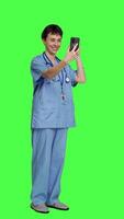 Side view Nurse answering videocall meeting connection on work break, using smartphone to connect with friends on online videoconference chat. Specialist in scrubs stands against greenscreen. Camera A. video