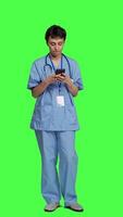 främre se hälsa specialist använder sig av smartphone till göra Viktig samtal, stående mot grönskärm bakgrund. sjuksköterska talande till patienter för kolla upp prov på telefon linje, kallelse människor till bekräfta. kamera a. video