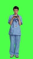 Front view Medical assistant playing mobile video games on smartphone app, taking a break from healthcare work. Nurse enjoys gaming tournament on phone, standing against greenscreen backdrop. Camera A.