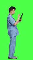 Profile Medical assistant browsing online webpages on tablet and texting, using social media apps to chat with people. Nurse navigates internet on gadget, standing against greenscreen backdrop. Camera A. video