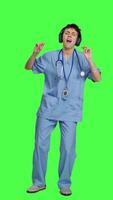 Front view Nurse having fun listening to music on wireless headphones, wearing scrubs and dancing around against greenscreen backdrop. Medical assistant showing groovy funky dance moves. Camera A. video