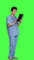 Side view Medical assistant browsing online webpages on tablet and texting, using social media apps to chat with people. Nurse navigates internet on gadget, standing against greenscreen backdrop. Camera A. video