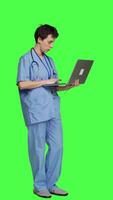 Side view Medical assistant browsing for healthcare treatments to help patients, using laptop against greenscreen backdrop. Young nurse navigates online websites an searches for health support. Camera A. video