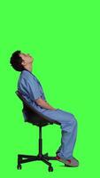 Profile Medical assistant is waiting for patients to attend exam appointments, feeling impatient while she sits in a chair. Nurse with stethoscope and scrubs waits for people against greenscreen. Camera A. video