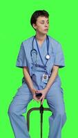 Front view Medical assistant waiting for someone while she sits on chair, feeling impatient and waits for patients to attend checkup appointments. Nurse in scrubs against greenscreen backdrop. Camera A. video