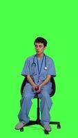 Front view Medical assistant is waiting for patients to attend exam appointments, feeling impatient while she sits in a chair. Nurse with stethoscope and scrubs waits for people against greenscreen. Camera A. video