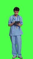 Front view Medical assistant browsing online webpages on tablet and texting, using social media apps to chat with people. Nurse navigates internet on gadget, standing against greenscreen backdrop. Camera A. video