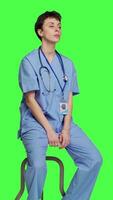 Side view Medical assistant waiting for someone while she sits on chair, feeling impatient and waits for patients to attend checkup appointments. Nurse in scrubs against greenscreen backdrop. Camera A. video