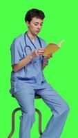 Side view Young nurse studying for the physician exam by reading book in studio, sitting on chair against greenscreen. Specialist enjoys healthcare industry literature, preparing to become a doctor. Camera A. video