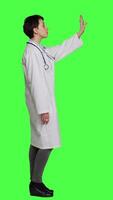 Profile Woman medic waving hello and asking people to come over at checkup, inviting patient to come closer and greets them. Skilled physician with white coat and stethoscope against greenscreen. Camera A. video