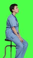 Profile Medical assistant waiting for someone while she sits on chair, feeling impatient and waits for patients to attend checkup appointments. Nurse in scrubs against greenscreen backdrop. Camera A. video