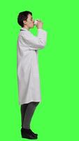 Profile Woman physician drinking a cup of coffee against greenscreen backdrop, taking a break from medical work. Specialist serving caffeine refreshment in studio, wearing a white coat. Camera A. video