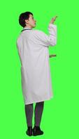 Back view Physician pointing to the left or right to create web commercial standing against greenscreen backdrop, doing a marketing advertisement in studio. Woman medic in white coat shows an ad. Camera A. video