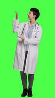 Front view Physician pointing to the left or right to create web commercial standing against greenscreen backdrop, doing a marketing advertisement in studio. Woman medic in white coat shows an ad. Camera A. video