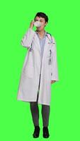 Front view Woman physician drinking a cup of coffee against greenscreen backdrop, taking a break from medical work. Specialist serving caffeine refreshment in studio, wearing a white coat. Camera A. video