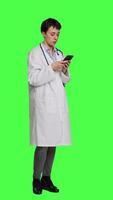 Side view Physician navigating on social media apps to text people, stands against greenscreen backdrop. Medic in white coat browsing websites online, making checkup appointments. Camera A. video