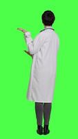 Back view Woman medic does web advertisement against greenscreen backdrop, pointing to left and right sides in studio. Doctor with white coat advertising something for a commercial. Camera A. video