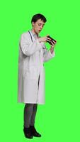 Side view General practitioner playing video games on mobile phone app, having fun with online gaming competition against greenscreen backdrop. Cheerful doctor relaxing with internet game. Camera A.