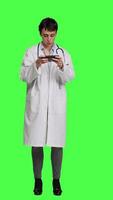 Front view General practitioner playing video games on mobile phone app, having fun with online gaming competition against greenscreen backdrop. Cheerful doctor relaxing with internet game. Camera A.