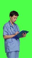 Side view Confident hospital nurse taking notes for the next medic appointment, standing over greenscreen backdrop. Medical assistant with uniform writing patient information on clipboard papers. Camera B. video