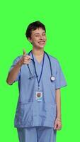 främre se glad sjuksköterska ger tummen upp tecken mot grönskärm bakgrund, som visar positiv gest med en tycka om. medicinsk assistent visar henne godkännande, rekommenderar något och varelse nöjd. kamera b. video